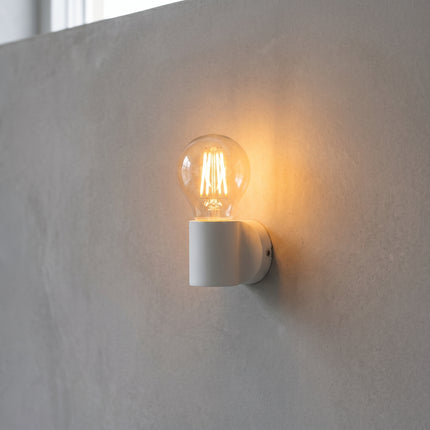Ampoule LED à filament E27 G45 allumée 3000K dans une applique murale blanche, halo chaud sur béton gris