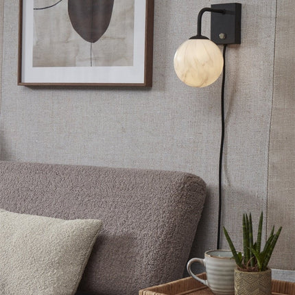 Applique murale Carrara globe en verre effet marbre blanc et bras noir mat, allumée près d'un canapé