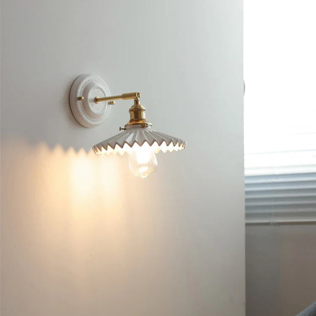 Applique murale en céramique blanche plissée et laiton allumée sur un mur clair de chambre