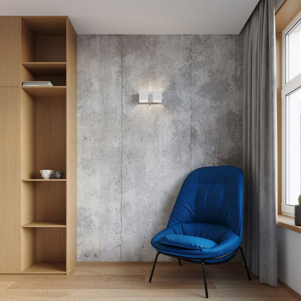 Applique Cube LED 2 feux blanche allumée sur mur béton, ambiance chaleureuse près d'un fauteuil bleu