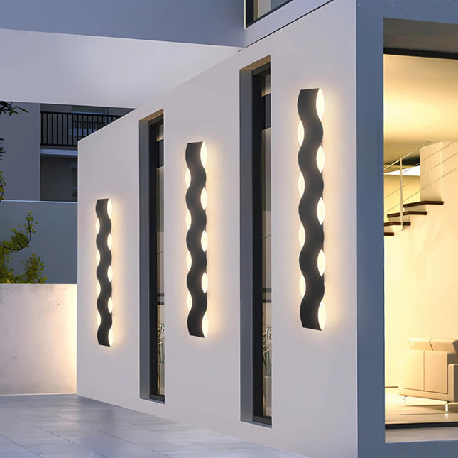 Applique extérieure LED ondulée en métal noir posée sur une façade blanche à la tombée de la nuit