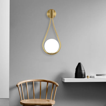 Applique murale goutte dorée à globe de verre opale sur un mur gris avec chaise en bois