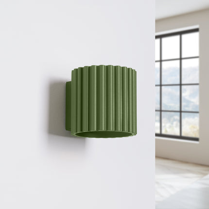 Applique Aura vert olive posée sur un mur blanc près d'une fenêtre dans un intérieur clair