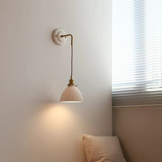 Applique murale en céramique blanche et cuivre, abat-jour galbé strié, allumée près d'une fenêtre dans une chambre cosy