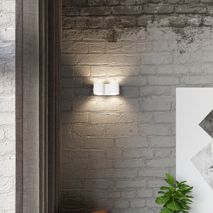 Applique cylindrique double blanche fixée sur un mur de brique, halos de lumière chaude au-dessus et en dessous.