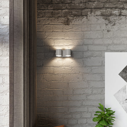 Applique cylindrique double grise fixée sur un mur de brique, halos de lumière chaude au-dessus et en dessous.