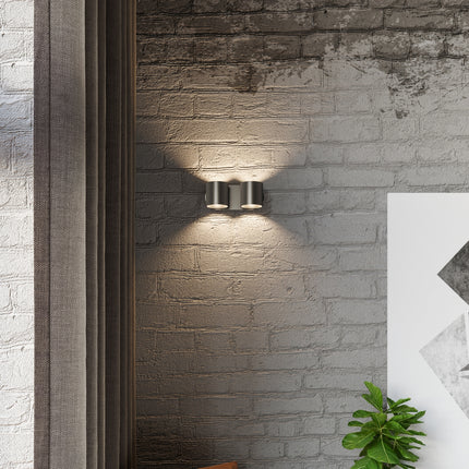 Applique cylindrique double noire fixée sur un mur de brique, halos de lumière chaude au-dessus et en dessous.