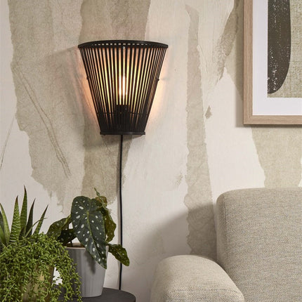 Applique murale Merapi en bambou noir tressé allumée dans un salon, à côté d'un fauteuil beige et de plantes vertes