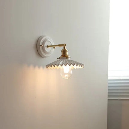 Détail de l applique murale orientable en céramique plissée et laiton posée sur un mur blanc, halo lumineux ambré
