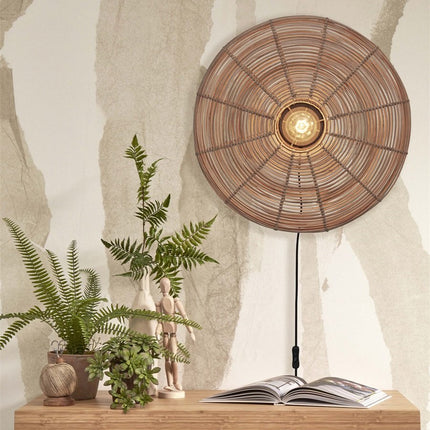 Tanami natural woven rattan wall light switched on against a textured wall, above a piece of furniture with plants