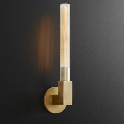 Applique murale tube en verre strié finition cuivre allumée sur un mur foncé, lumière chaude