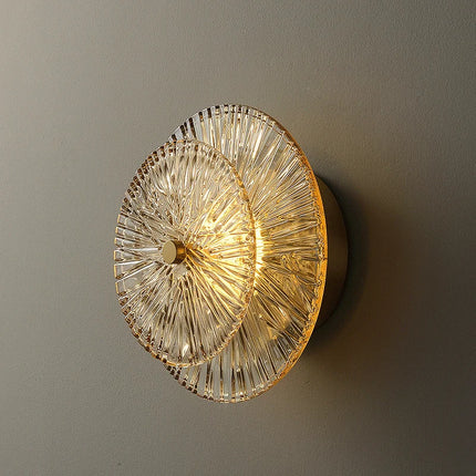 Applique murale en verre strié doré à motif soleil, allumée en lumière blanc chaud sur un mur gris