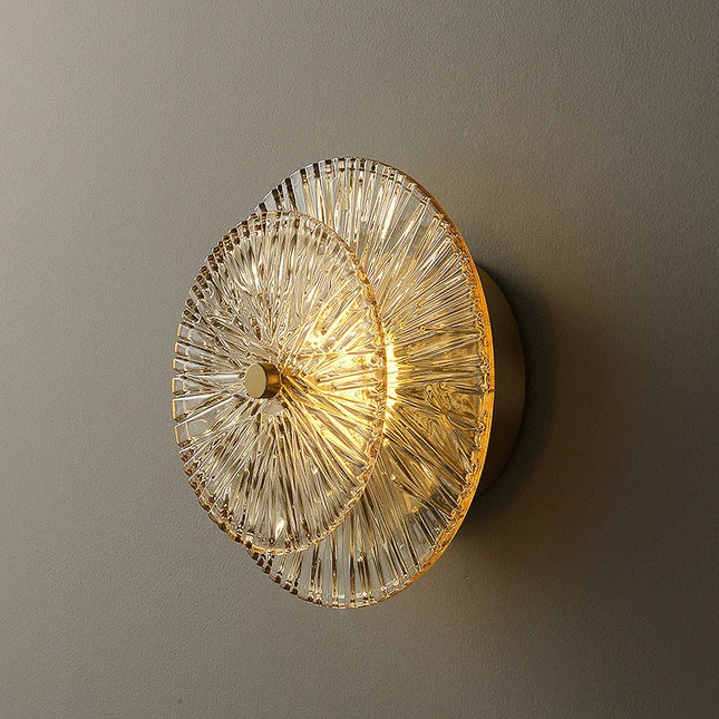 Applique murale en verre strié doré à motif soleil, allumée en lumière blanc chaud sur un mur gris