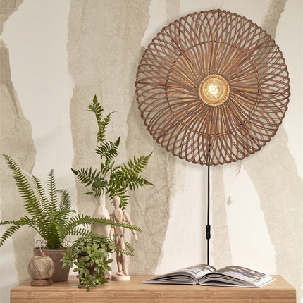 Zanzibar natural woven rattan wall light switched on above a wooden cabinet with green plants