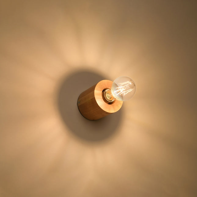 Applique en bois naturel allumée au mur, halo de lumière chaude autour du cylindre.
