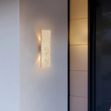 Applique en travertin posée sur une façade moderne près d'une baie vitrée, halo chaud le soir