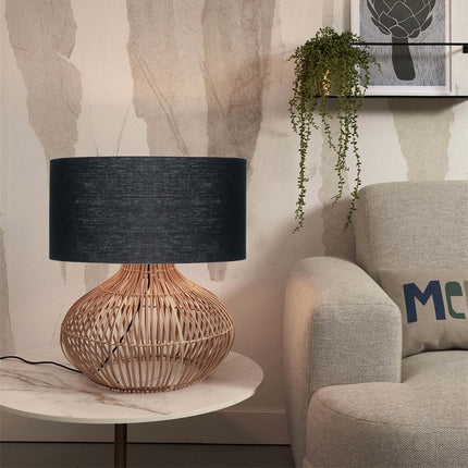 Lampe à poser Kalahari en rotin tressé et abat-jour lin noir posée sur une table près d'un fauteuil