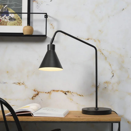 Lampe à poser Lyon en fer noir posée sur un bureau en bois, abat-jour conique orienté vers un livre ouvert