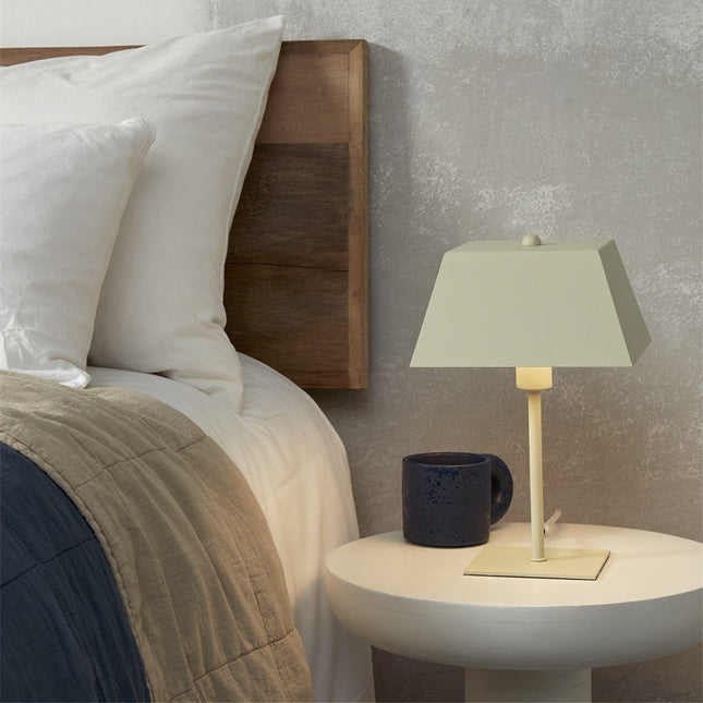 Lampe à poser au design carré posée sur une table de chevet ronde, à côté d'un lit