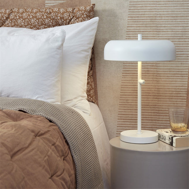 Lampe à poser Porto fer blanc allumée sur une table de chevet dans une chambre chaleureuse
