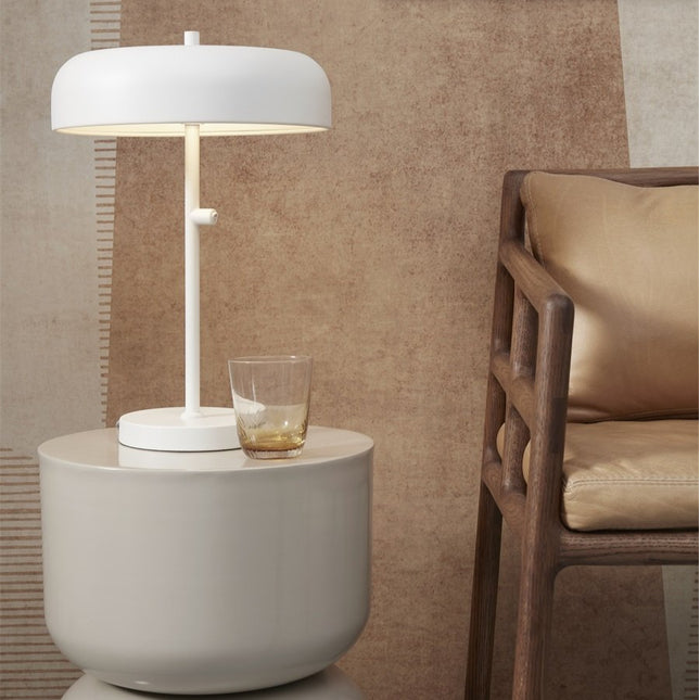 Lampe à poser Porto en fer blanc sur une table de chevet ronde, à côté d'un fauteuil en bois