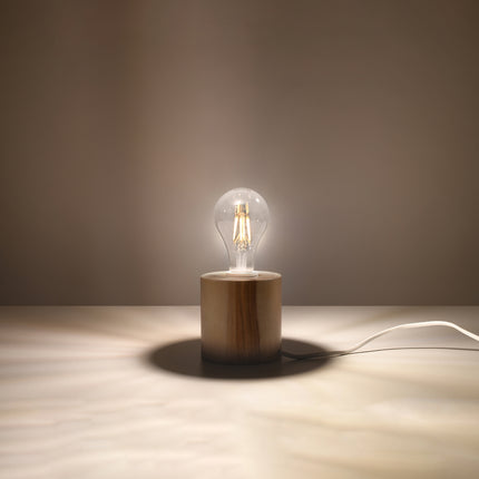 Lampe à poser en bois naturel allumée sur une table, lumière chaude d'ambiance et câble apparent.