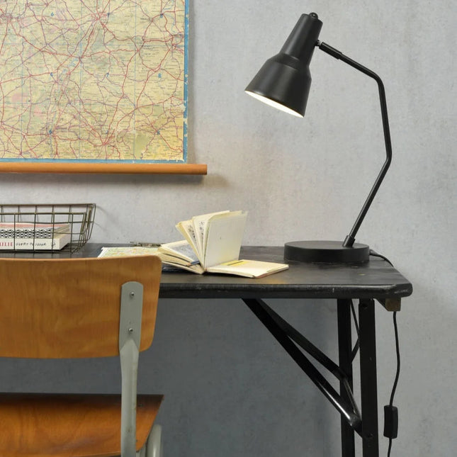 Lampe à poser Valencia en fer noir posée sur un bureau industriel, bras articulé et lumière chaude dirigée
