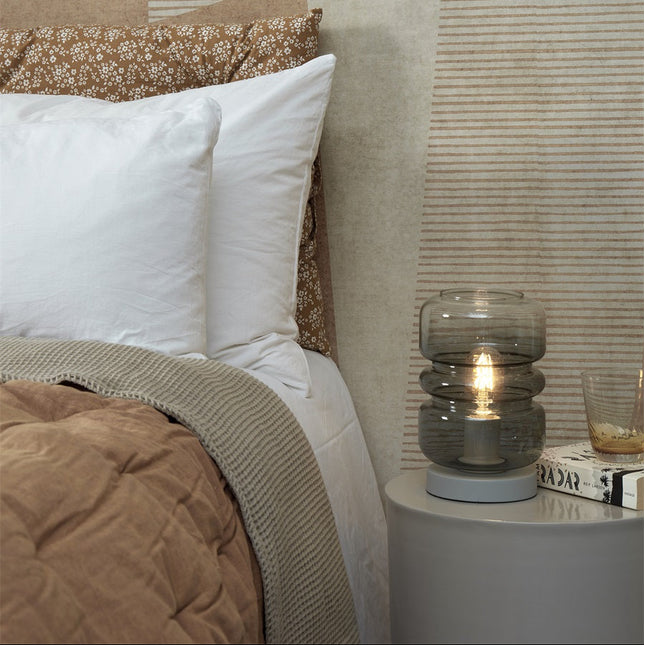 Lampe à poser Verona en verre fumé gris posée sur une table de chevet ronde