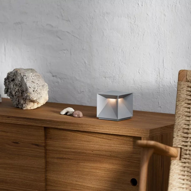 Lampe à poser LED Nutalis en aluminium gris brume posée sur un meuble en bois, forme cubique biseautée, ambiance chaleureuse