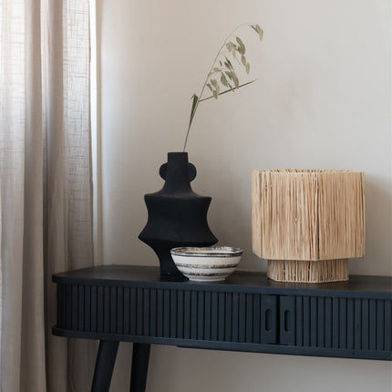Lampe Panama raphia naturel sur une console noire, à côté d'un vase sculptural et d'un bol en céramique