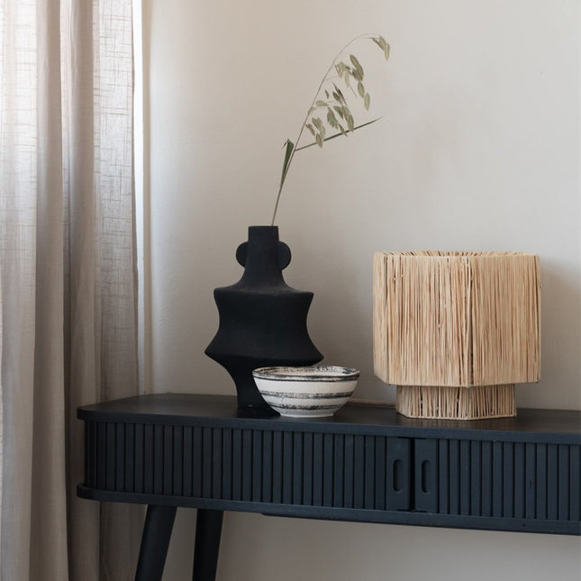 Lampe Panama raphia naturel sur une console noire, à côté d'un vase sculptural et d'un bol en céramique