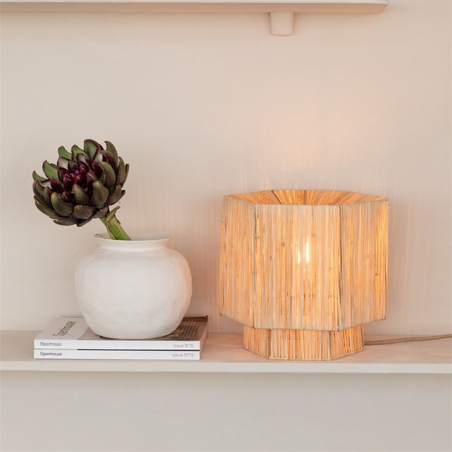 Lampe à poser Panama en raphia naturel tressé, abat-jour hexagonal, posée sur une étagère près d'un vase blanc