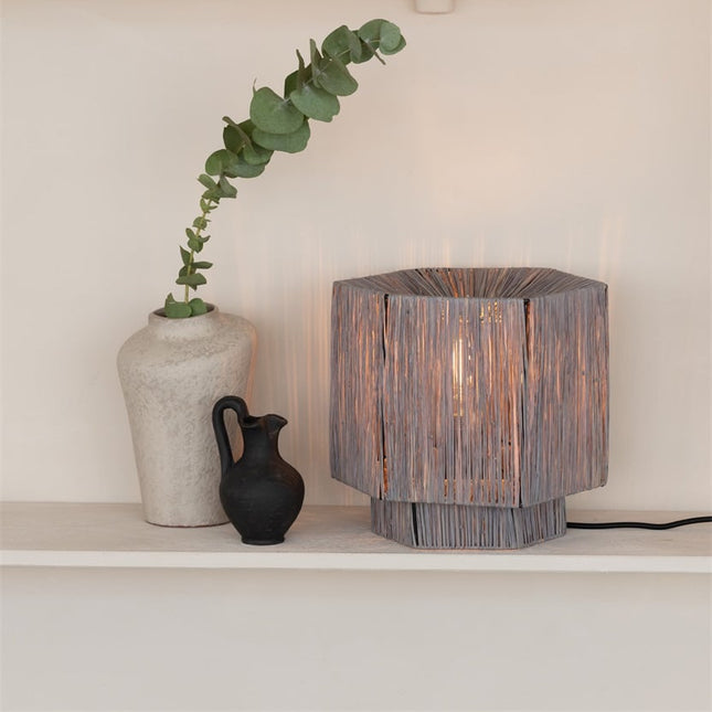 Lampe à poser Panama allumée sur une étagère, à côté d'un vase en céramique et d'une plante