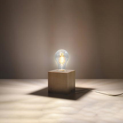 Lampe à poser cube en bois naturel allumée, ampoule filament chaude posée sur le cube, ombre douce.