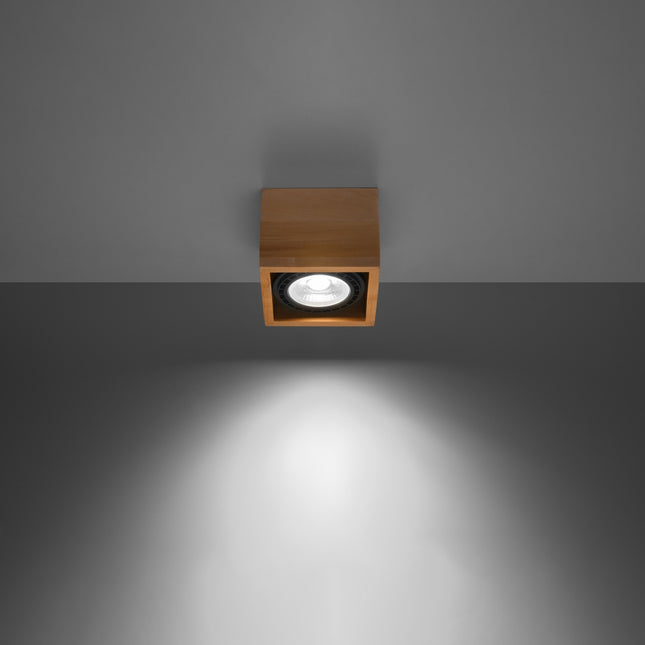 Plafonnier spot cube bois naturel projetant un faisceau lumineux sur un mur gris