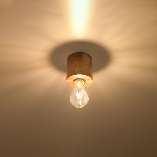 Plafonnier en bois naturel allumé au plafond, lumière chaude diffusée en halo autour du cylindre.