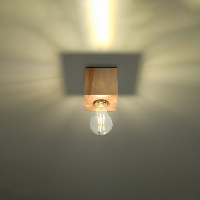 Plafonnier cube en bois naturel allumé, halo chaud diffusé par l'ampoule filament E27 sous le plafond.