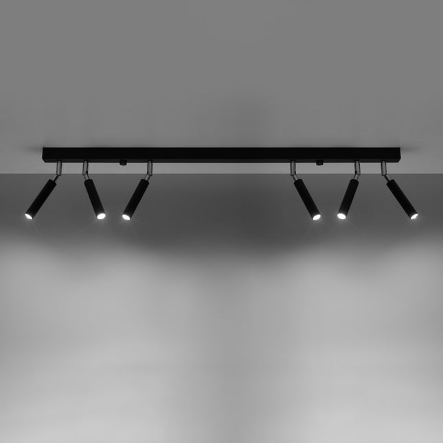 Rampe de 6 spots acier noir allumee projetant ses faisceaux sur un mur clair