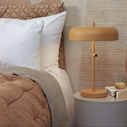Lampe à poser Porto moutarde sur une table de chevet ronde, dans une chambre lumineuse