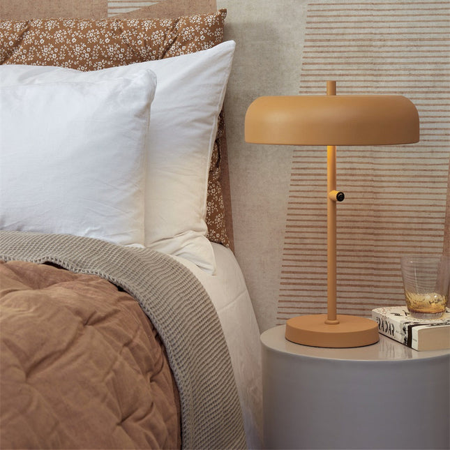 Lampe à poser Porto moutarde sur une table de chevet ronde, dans une chambre lumineuse